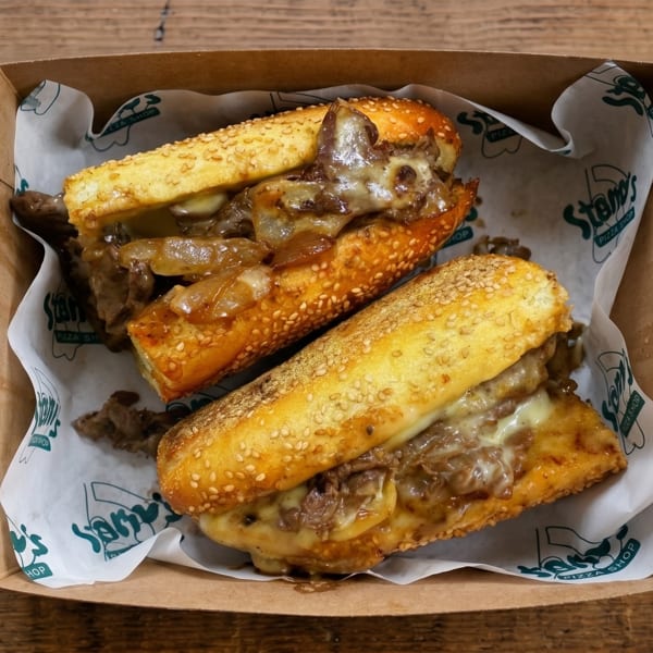 Two cheesy steak and onion sandwiches in a cardboard box with branded paper lining.