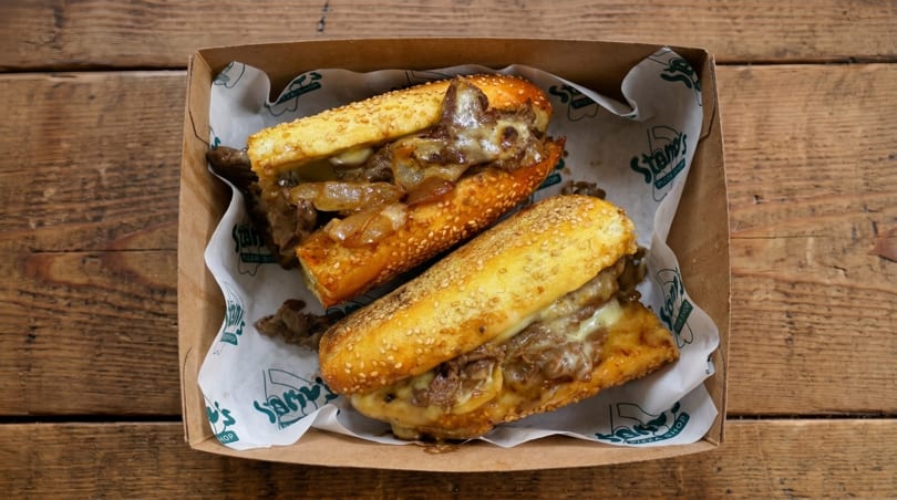 Two cheesy steak and onion sandwiches in a cardboard box with branded paper lining.