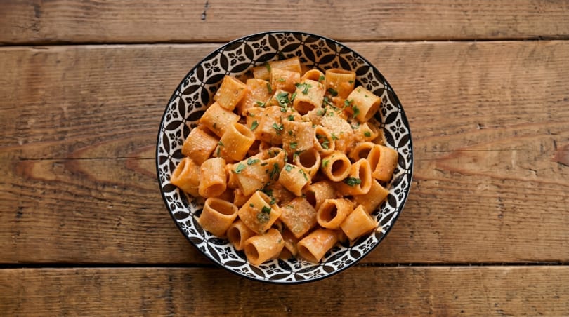 A black and white patterned bowl of rigatoni pasta with red sauce and parsley on a wooden table.